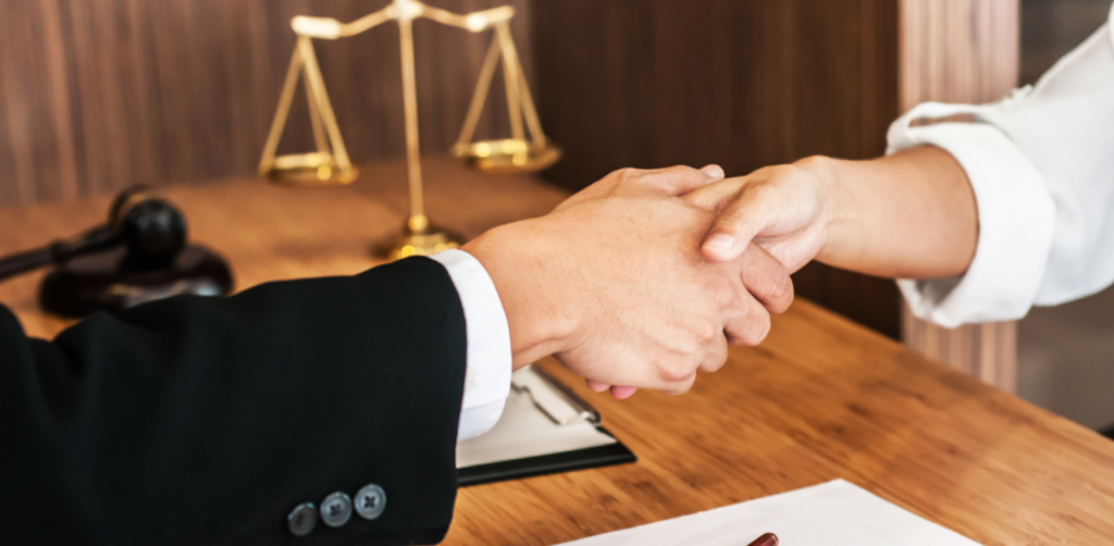 Two people shaking hands across a desk in an office, with scales of justice and a gavel visible in the background