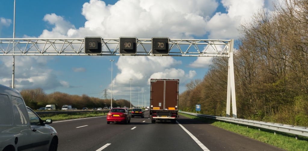 Highway scene with multiple cars and a truck under overhead speed limit signs displaying "90", "90", and "70" against a partly cloudy sky