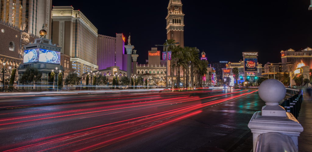 Vibrant nighttime view of the Las Vegas Strip, with illuminated hotels, palm trees, and red light trails from moving vehicles on the road