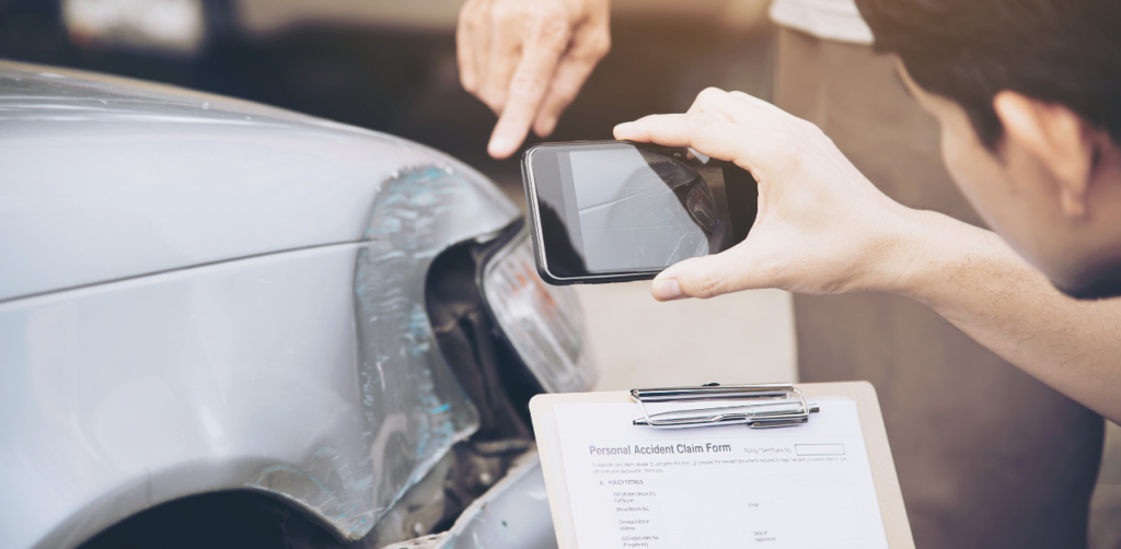 A person taking a photo of a damaged car bumper with a smartphone while another person points at the damage; a claim form is visible on a clipboard