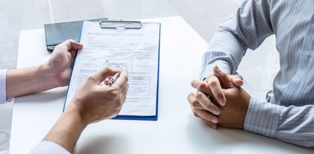 Two people discussing a document on a clipboard; one person holds a pen while the other sits with clasped hands