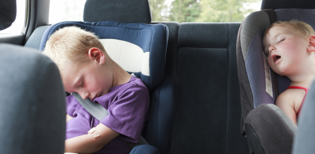 Two children asleep in car seats in the backseat of a car; one child in a purple shirt, the other with blonde hair wearing a red tank top