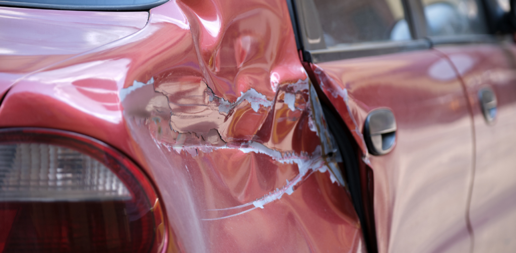 A close-up of the rear side of a red car with visible dents and scratches from an accident