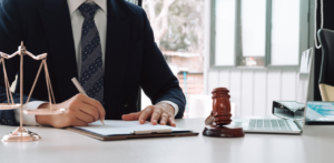 A lawyer in a suit writing on a document at a desk, with a gavel, scales of justice, and a laptop nearby
