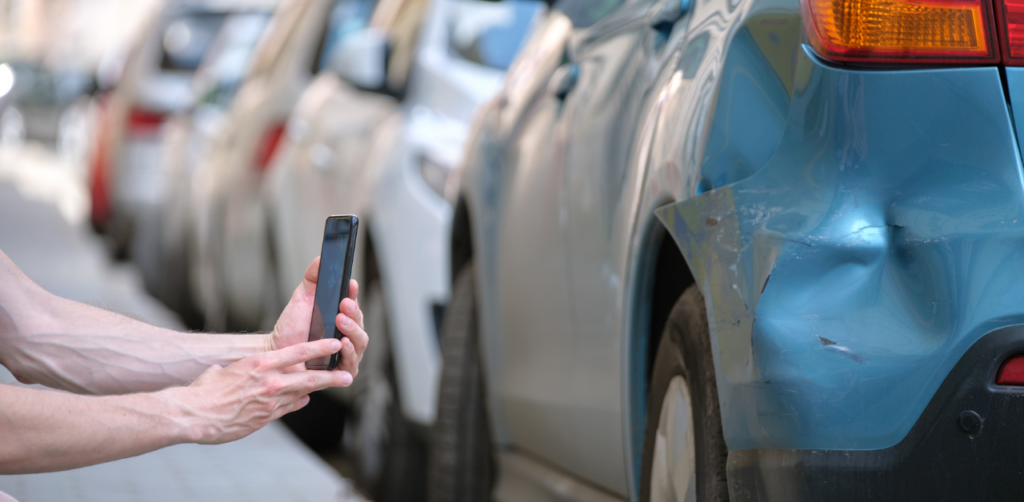 A person photographing damage to the rear of a blue car, parked alongside other vehicles