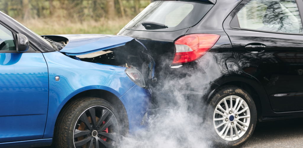 Blue car with a crumpled hood has rear-ended a black car, with smoke rising from the impact site on a roadside