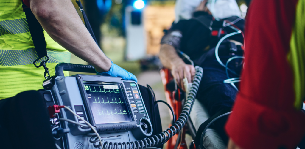 Emergency responders tend to a patient on a stretcher while monitoring an ECG display on medical equipment in an outdoor setting