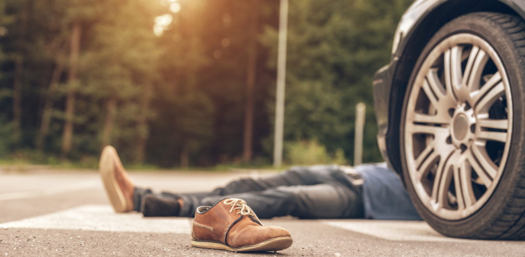 Shoe lies on the ground near an injured person lying next to a car on a crosswalk, with a forested background and sunlight filtering through