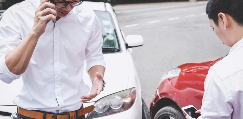Two men discussing a car accident, with a red and a white car visibly damaged in the background