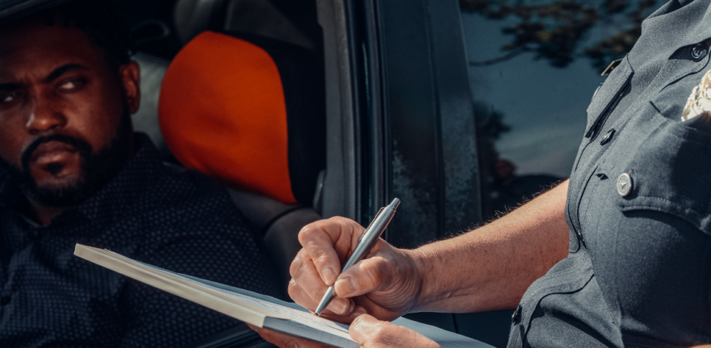 A police officer is writing a ticket next to a car window while the driver, a man, looks out from inside the vehicle