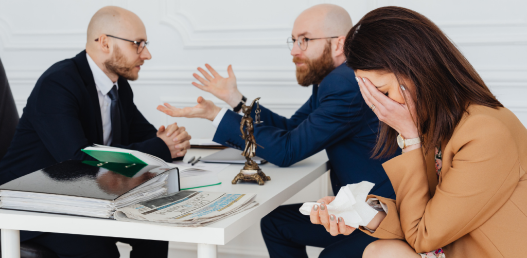 Two men in suits are in a heated discussion across a table, while a woman sits to the side, covering her face with her hand and holding a tissue