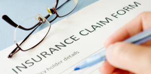 A close-up of a hand filling out an insurance claim form with a pen, with reading glasses placed on top of the form