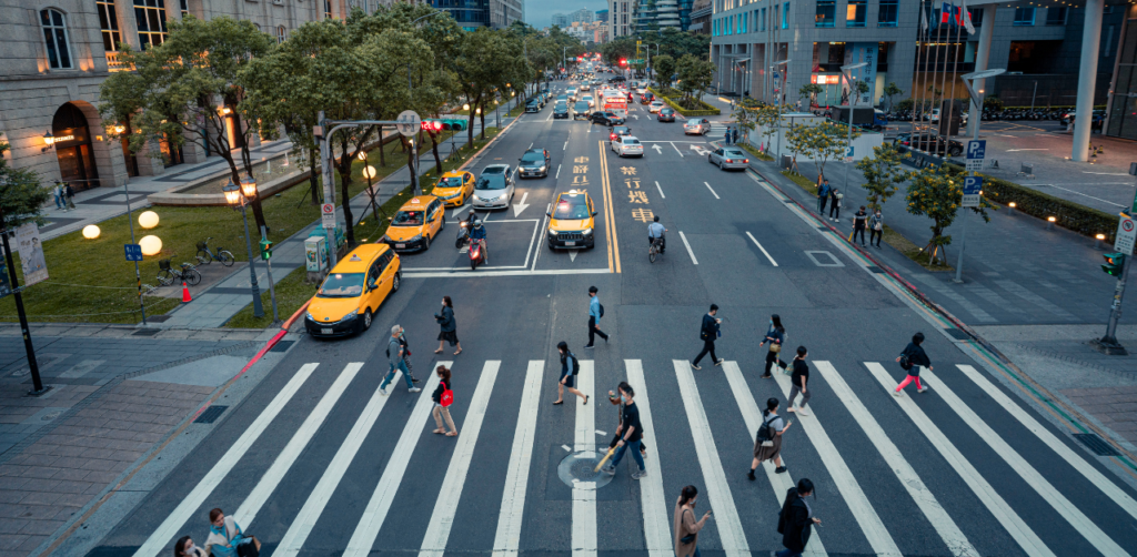 Aerial view of a busy city intersection with multiple pedestrians crossing on wide zebra crosswalks, while cars and taxis wait at the traffic light