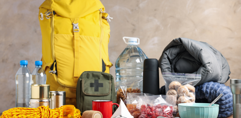 Emergency supplies, including a yellow backpack, water bottles, canned food, a first aid kit, and other essentials, arranged on a table