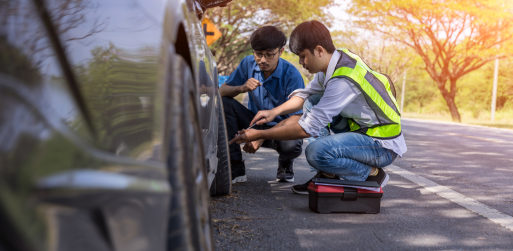 Two men kneeling next to a car on the side of the road, examining a tire with tools, one wearing a reflective safety vest