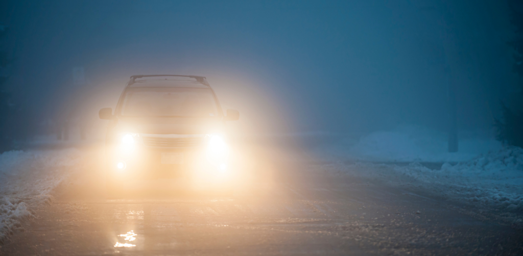 A car with headlights on driving through thick fog on a snowy road at night