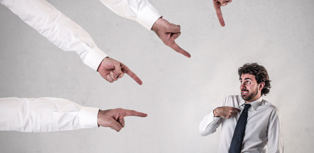 A nervous man in a suit being pointed at by multiple hands from off-frame, conveying a sense of accusation or blame