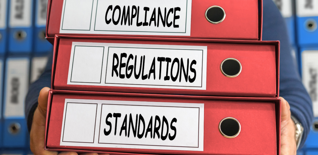 A person holding three red binders stacked, labeled "COMPLIANCE," "REGULATIONS," and "STANDARDS."