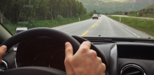 View from inside a car of a driver's hands on the steering wheel, with a clear road and green landscape ahead
