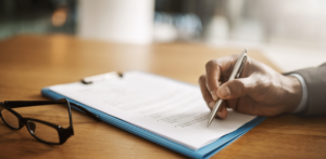 A person signing a document on a clipboard, with reading glasses resting on the table