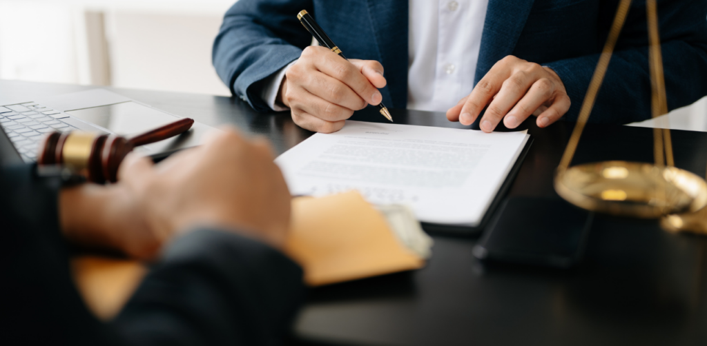 Two people in business attire signing legal documents at a desk with a gavel, scales, and a laptop, indicating a legal or contractual agreement