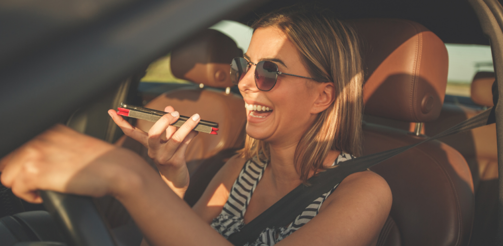Smiling woman in sunglasses driving a car while holding a smartphone, seemingly using voice commands