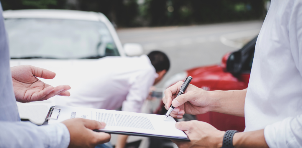 Two individuals exchanging information on a clipboard after a car accident, with damaged vehicles in the background