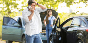 A stressed man calls for help after a car accident, with a concerned woman standing by