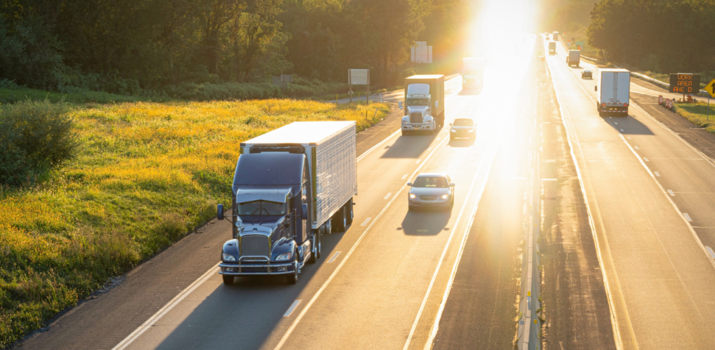 A highway with trucks and cars driving under a bright sun, bordered by green fields and trees, with sunlight streaming across the road