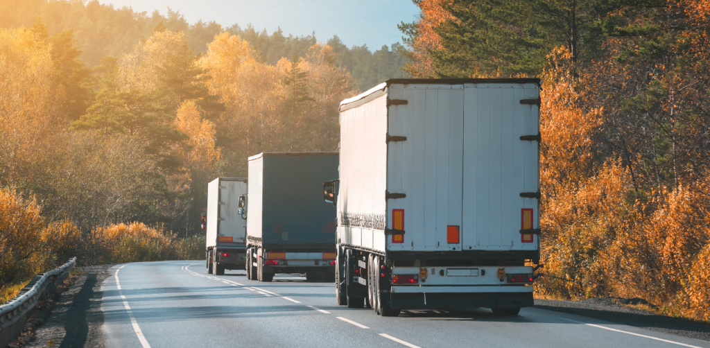 A line of cargo trucks driving on a scenic road surrounded by autumn-colored trees and soft morning light