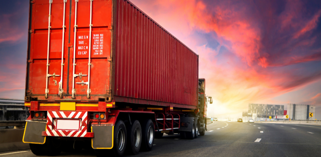 A red cargo truck with a shipping container driving on a highway under a vibrant sunset sky, with urban buildings in the distance