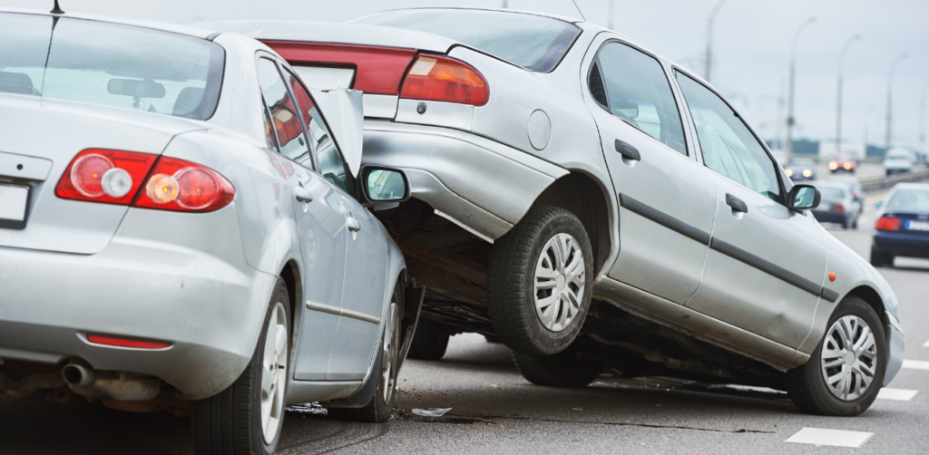 A silver car rear-ends another silver car, with the front of one vehicle lifted onto the back of the other on a busy roadway