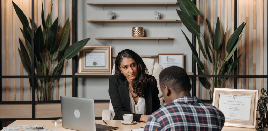 A lawyer attentively listening to a client across a desk in an office, with framed certificates in the background