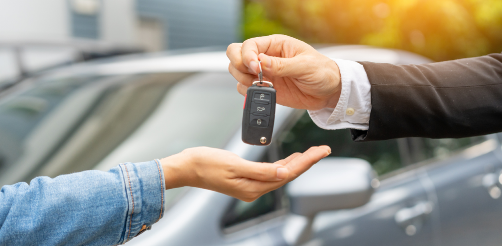 A person handing over car keys to another individual, with a car in the background, indicating a car sale or rental