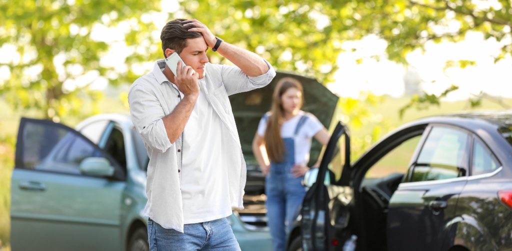 A man on the phone looking distressed after a car accident, with two cars and a woman in the background