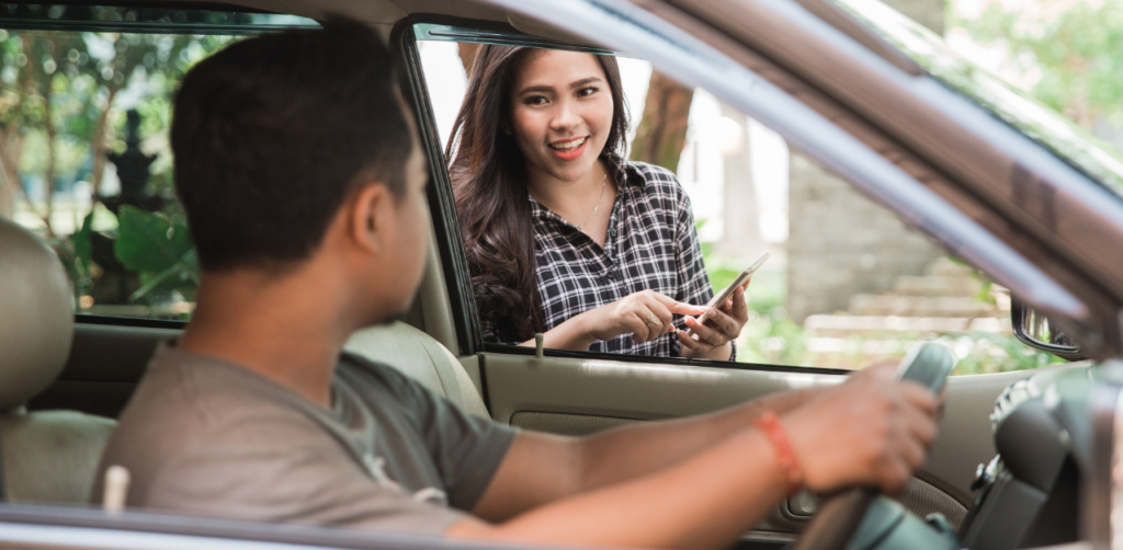 A man inside a car looking at a woman outside the car, who is holding a phone and smiling