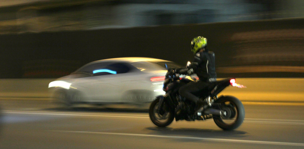 A motorcyclist in a black outfit and yellow helmet rides at high speed next to a blurry white car on a city street at night