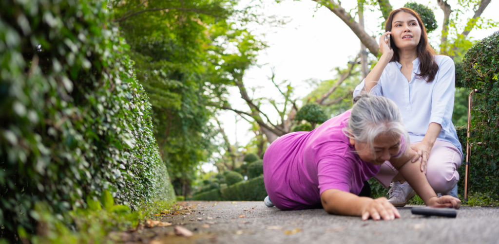 Older woman lying on the ground in a garden, reaching out while a younger woman calls for help on a phone