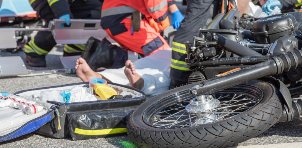 Paramedics and firefighters attend to an injured person lying on the ground near a motorcycle, with medical equipment scattered nearby
