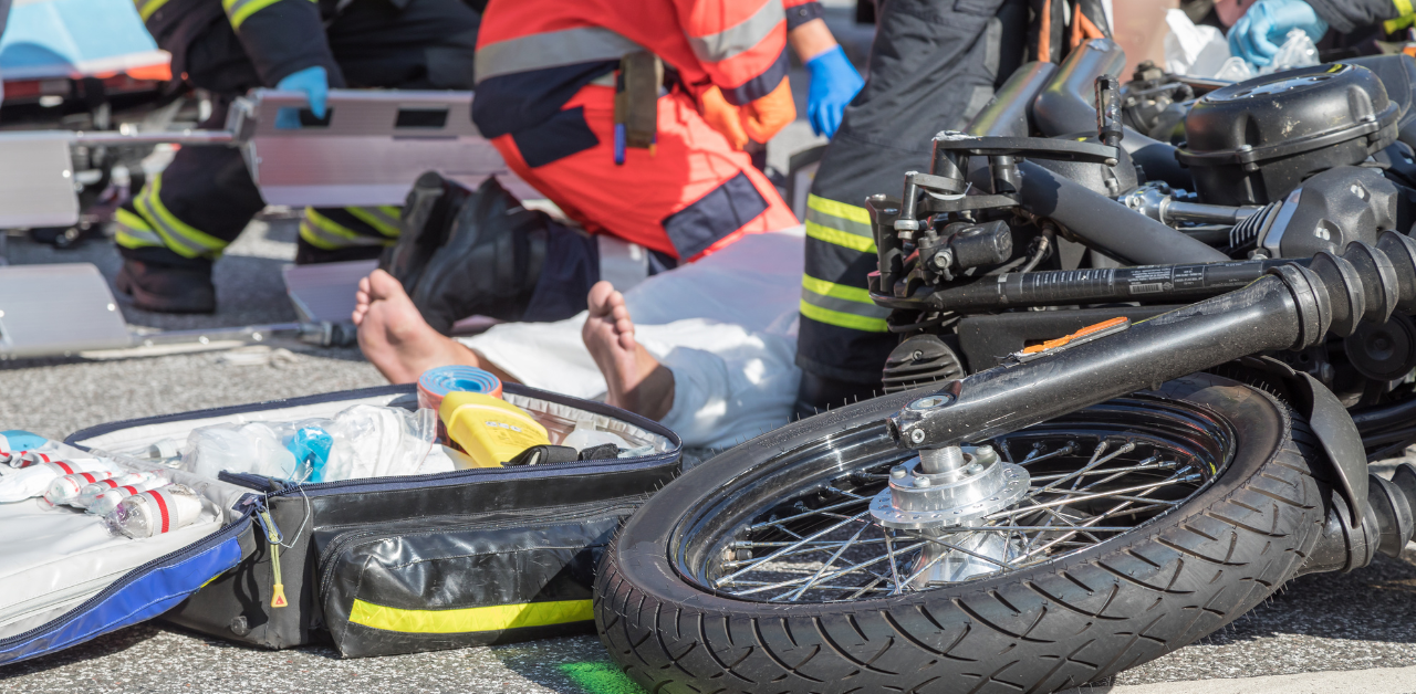 Paramedics and firefighters attend to an injured person lying on the ground near a motorcycle, with medical equipment scattered nearby