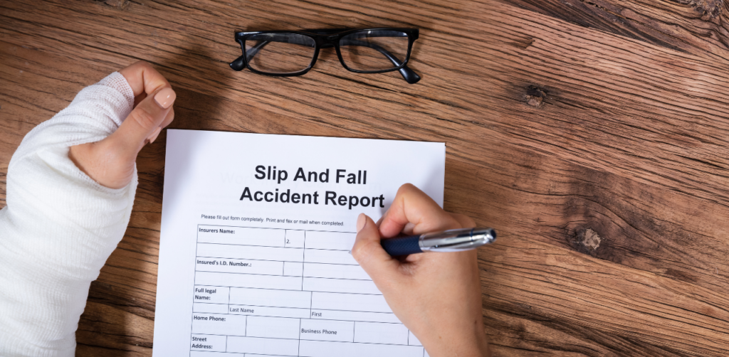 Person with a bandaged hand filling out a 'Slip and Fall Accident Report' form on a wooden table, with glasses nearby