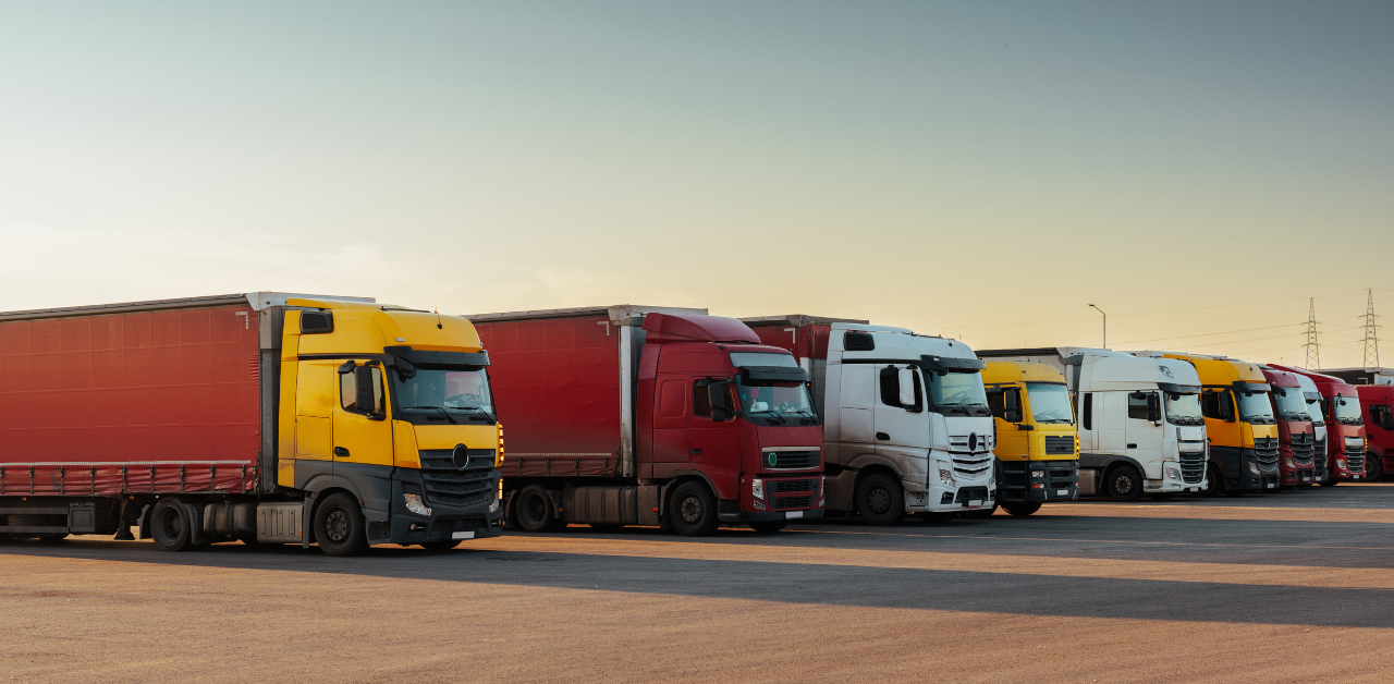 A row of semi-trucks with various colors, including yellow, red, and white, parked in a lot under a clear sky at sunset