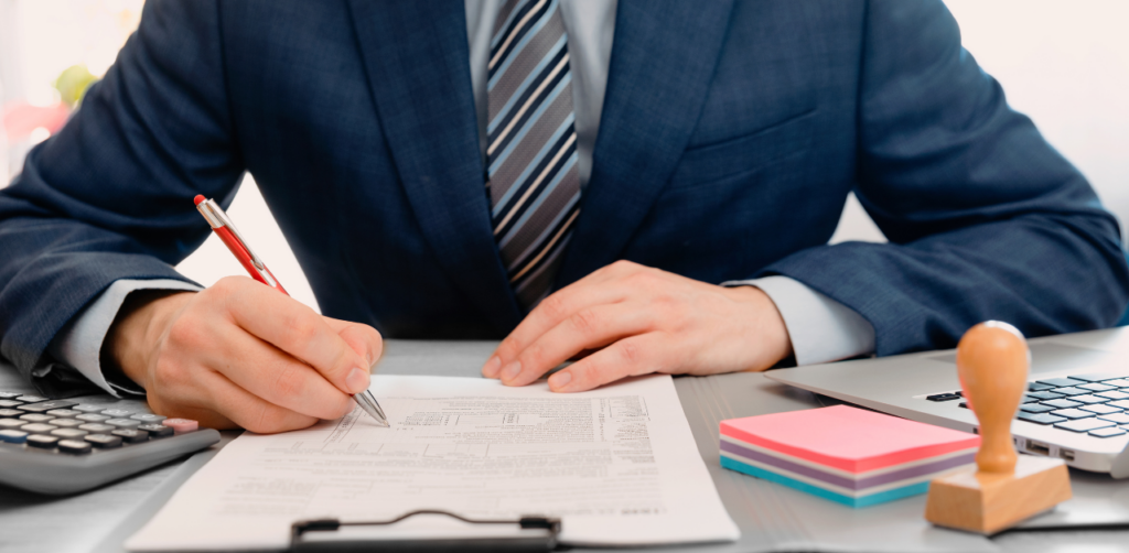 A man in a suit is filling out a form on a clipboard at a desk with a calculator, stack of sticky notes, and a rubber stamp beside him