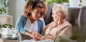 A younger woman sitting beside an elderly woman in a chair, holding her hand and speaking in a caring manner, conveying a sense of support or companionship