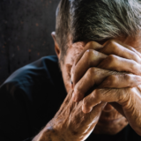 An elderly man sitting with his head down, hands clasped over his forehead, conveying a sense of sadness or contemplation