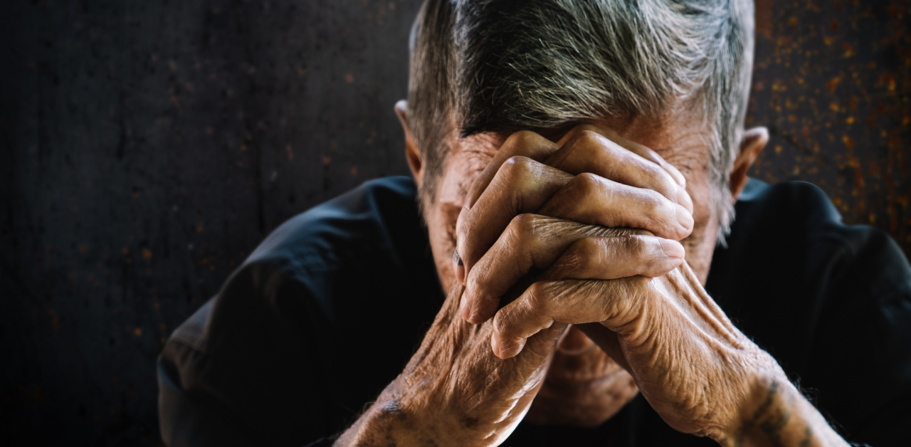 An elderly man sitting with his head down, hands clasped over his forehead, conveying a sense of sadness or contemplation