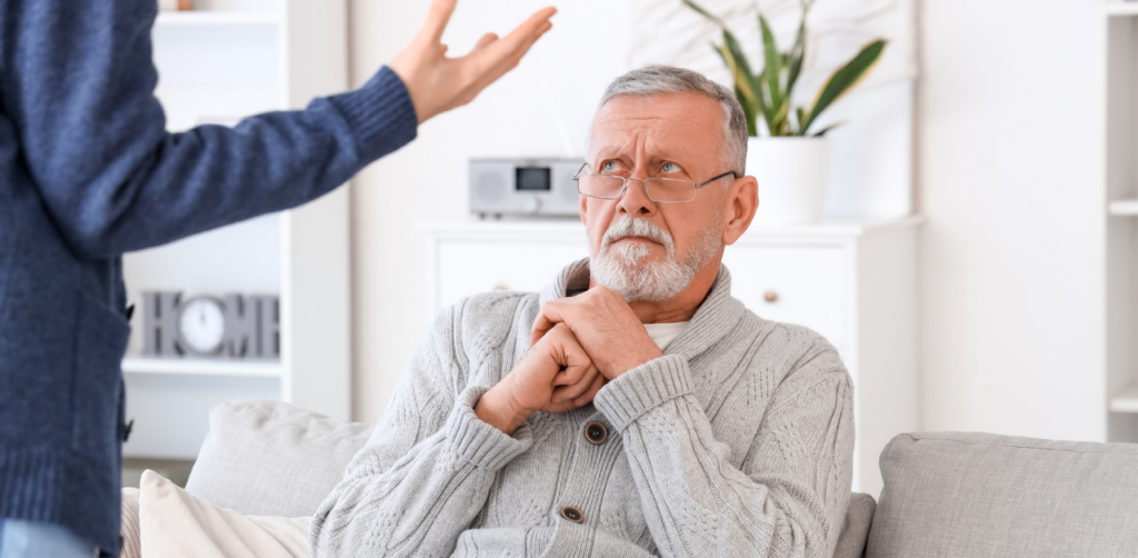 An older man sitting on a couch, looking up with a concerned expression while a person gestures towards him, suggesting a tense conversation