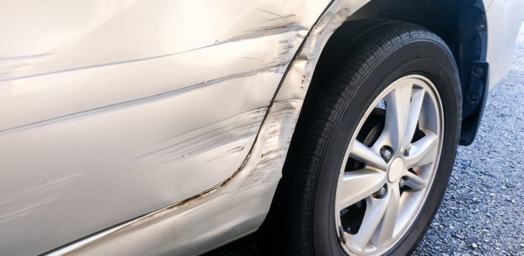 Close-up of a silver car with visible scratches and dents along the side, near the rear wheel