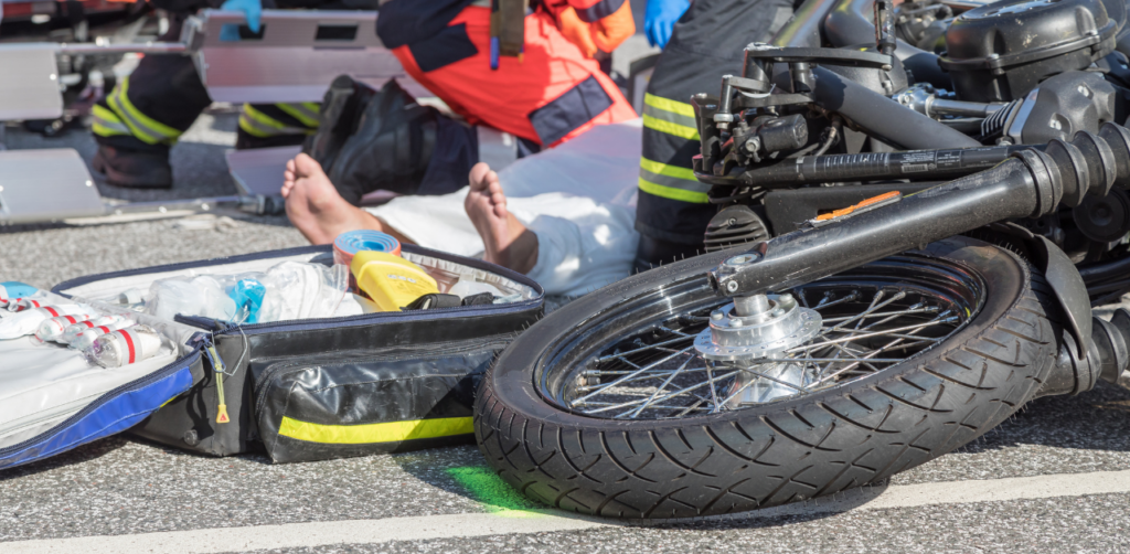 Emergency responders attend to a motorcyclist on the ground after an accident, with a motorcycle and medical kit visible in the foreground