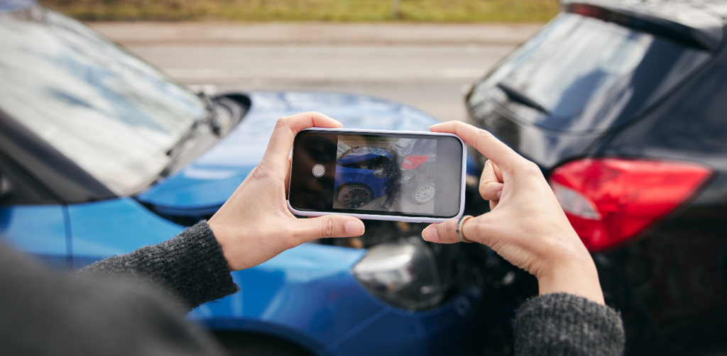 Hands holding a smartphone taking a photo of a car accident scene, with two damaged cars visible in the phone’s screen